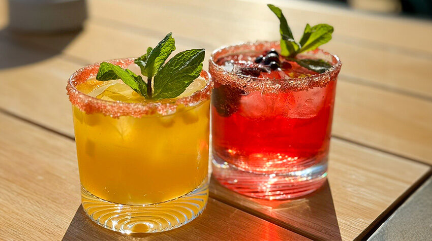 Ein gelber und ein roter Cocktail, die auf einem Holztisch im Sonnenlicht stehen und sich als Sommergetränke präsentieren.