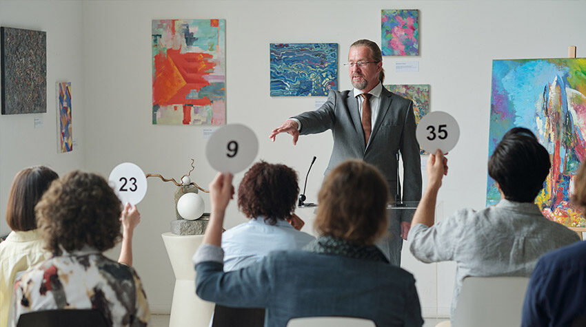 Auktionator bei Kunstauktion, der auf Bieter zeigt, die nummerierte Schilder hochhalten. ©Adobe Stock, pressmaster