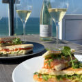 Zwei belegte Fischbrötchen mit Fisch, Salat und Kräutern auf Tellern sowie zwei Gläser Secco auf einem Holztisch mit Blick auf die Ostsee.