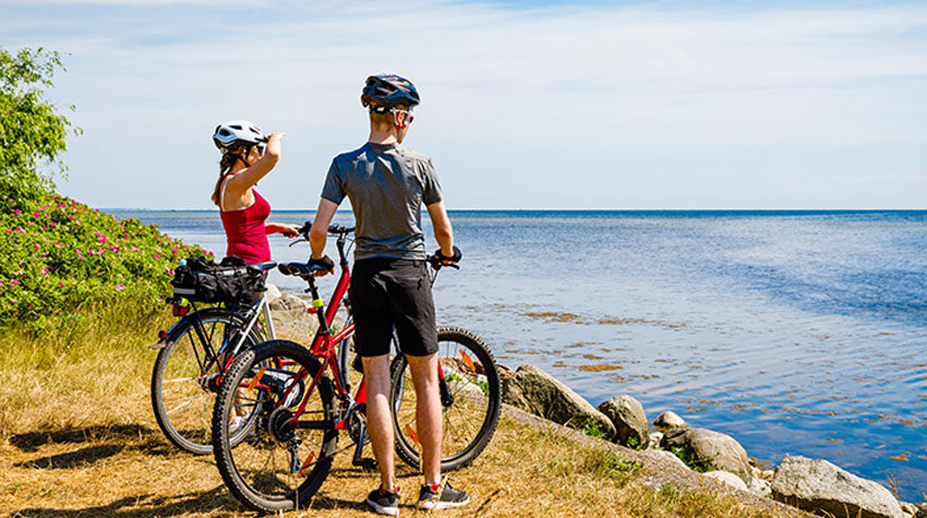 Radfahrerin und Radfahrer mit Helmen blicken an einem Sommertag aufs ruhige Meer an der Ostseeküste.