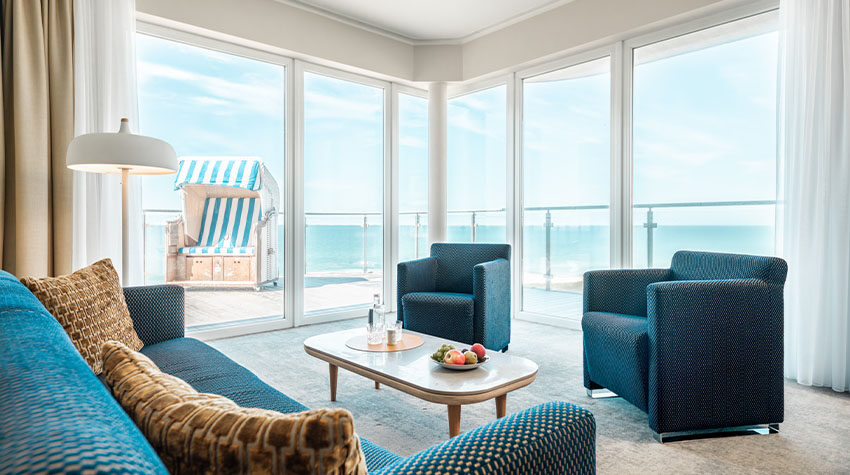 Helle Suite mit blauem Sofa, gemütlicher Leseecke und großem Balkon mit Strandkorb und Meerblick. im Hotel.