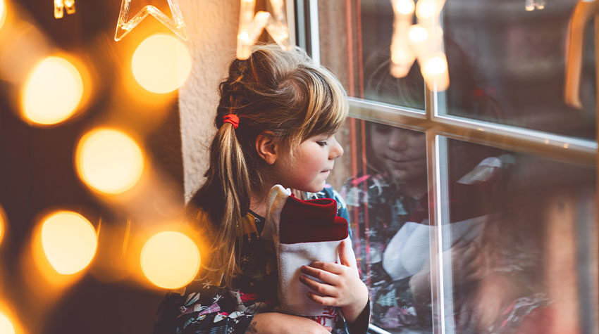 Ein Mädchen sitzt am beleuchteten Fenster, in der Hand hält sie einen Nikolausstiefel.