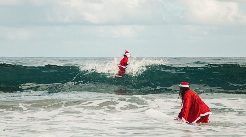 Zwei Personen im Weihnachtsmannkostüm surfen auf dem Meer.