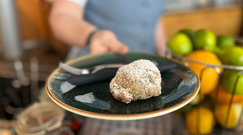Eine Person hält einen Teller mit einem einzelnen, mit Puderzucker bestäubten Stück Stollenkonfekt; daneben liegen Messer und Gabel.