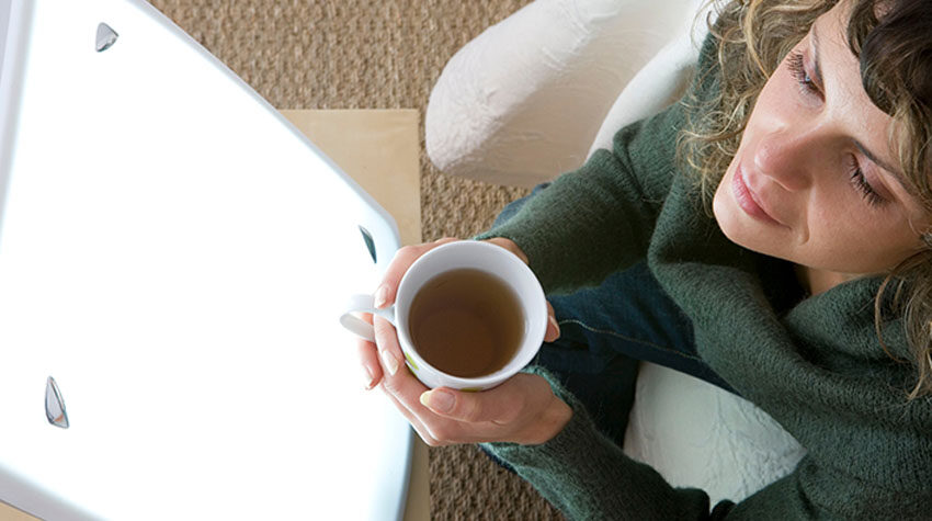 Frau sitzt entspannt mit einer Tasse in der Hand vor einer Lichttherapielampe.