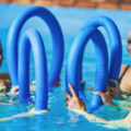 Vier Frauen trainieren beim Aquafitness im Pool mit blauen Schwimmnudeln.